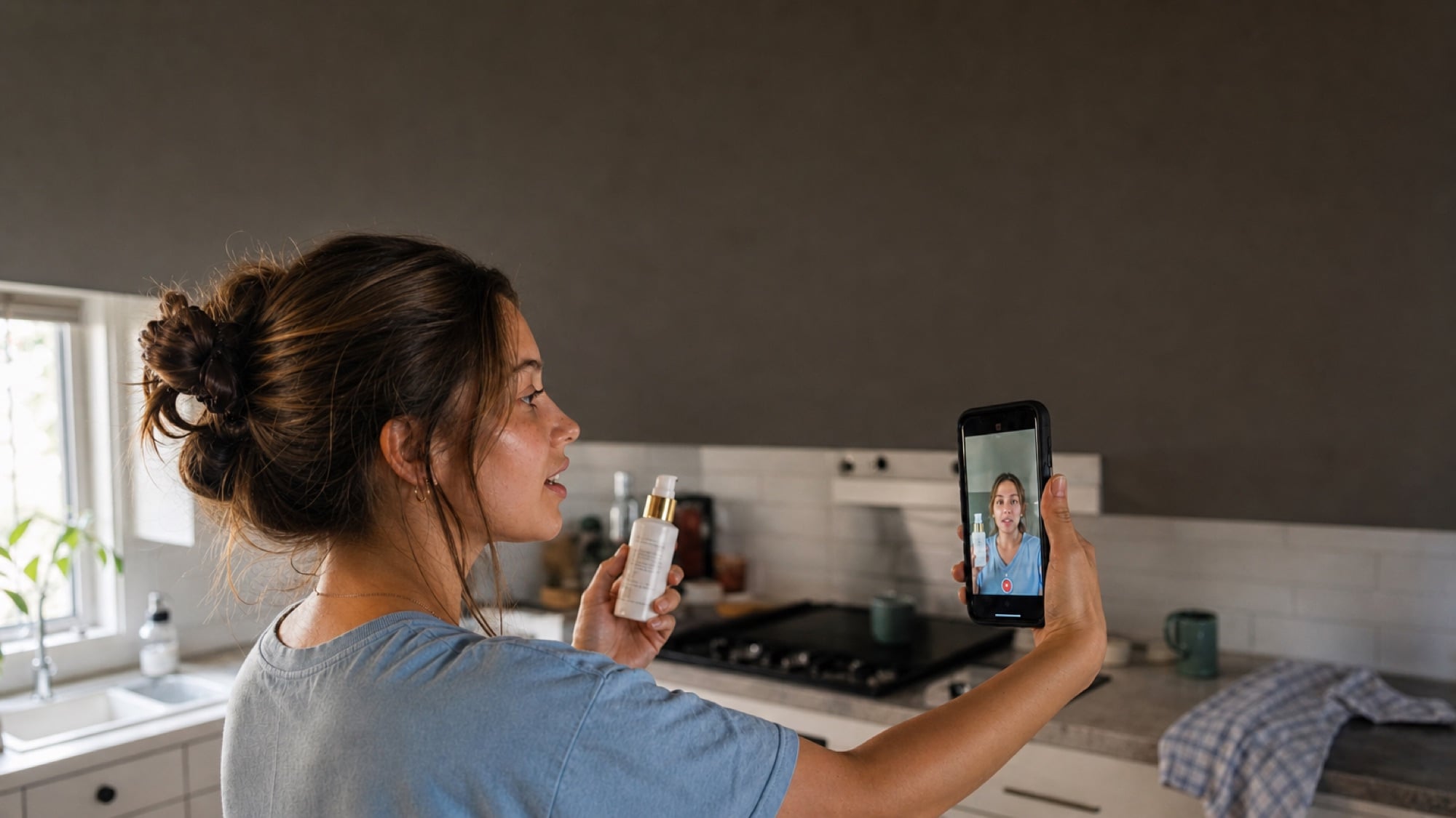 A creator filming herself on a phone in a kitchen, holding a skincare bottle to camera.